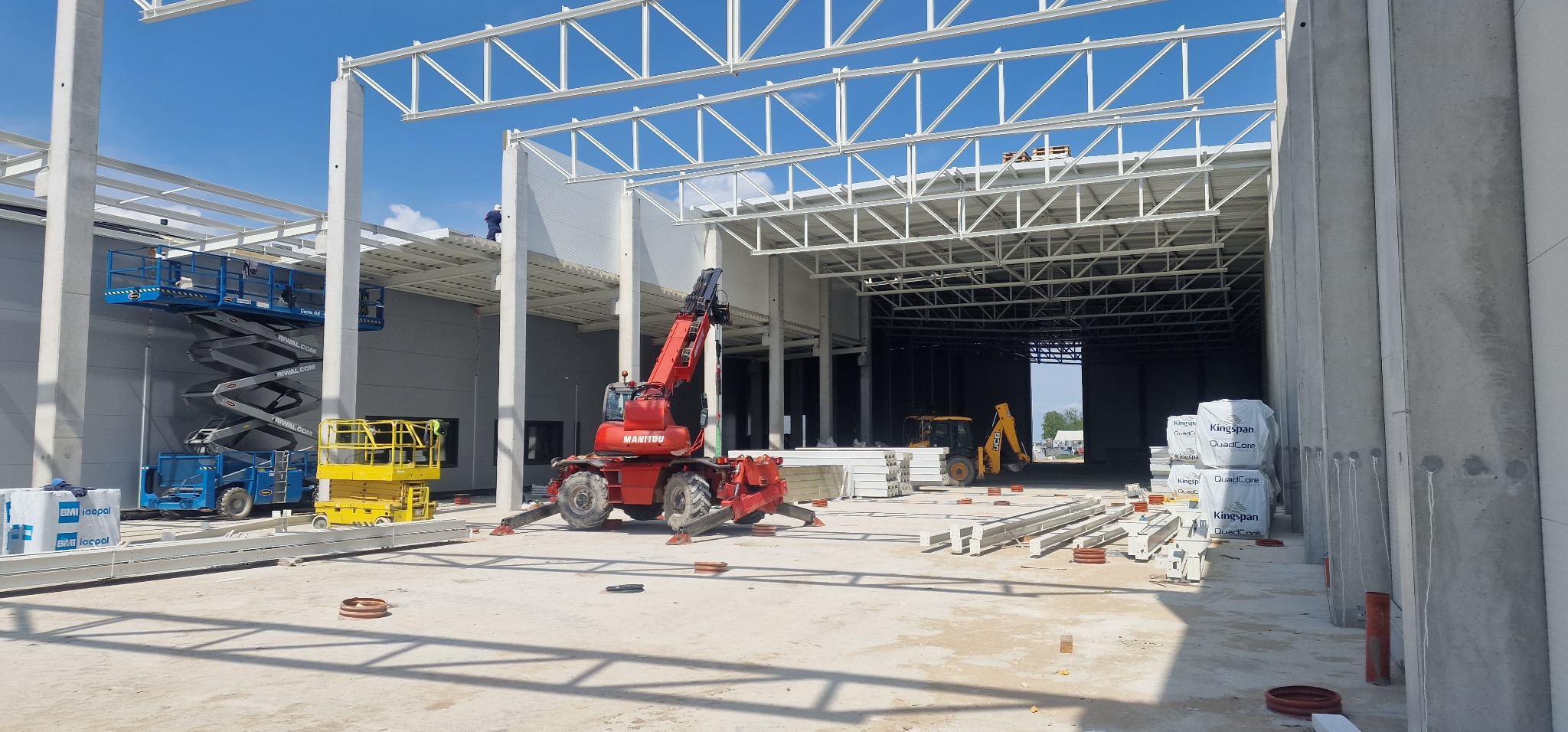 Montaż konstrukcji stalowej hali z użyciem dźwigu Manitou. W tle widoczne podnośniki koszowe i koparka. Elementy konstrukcyjne i płyty warstwowe na placu budowy.