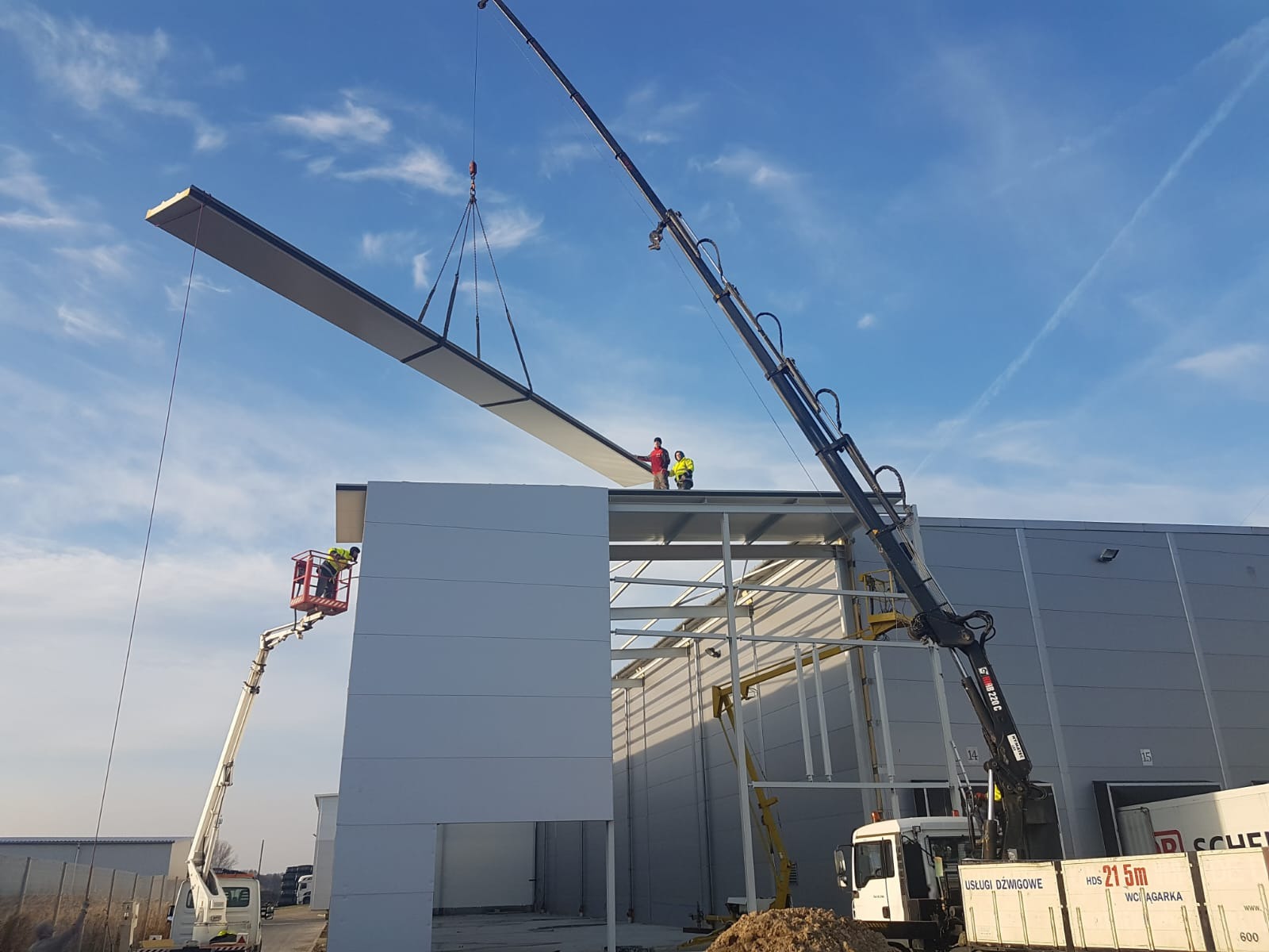 Montaż płyty warstwowej na budowie hali. Dwóch pracowników na dachu, jeden w podnośniku koszowym. Dźwig podnosi płytę. Błękitne niebo w tle.