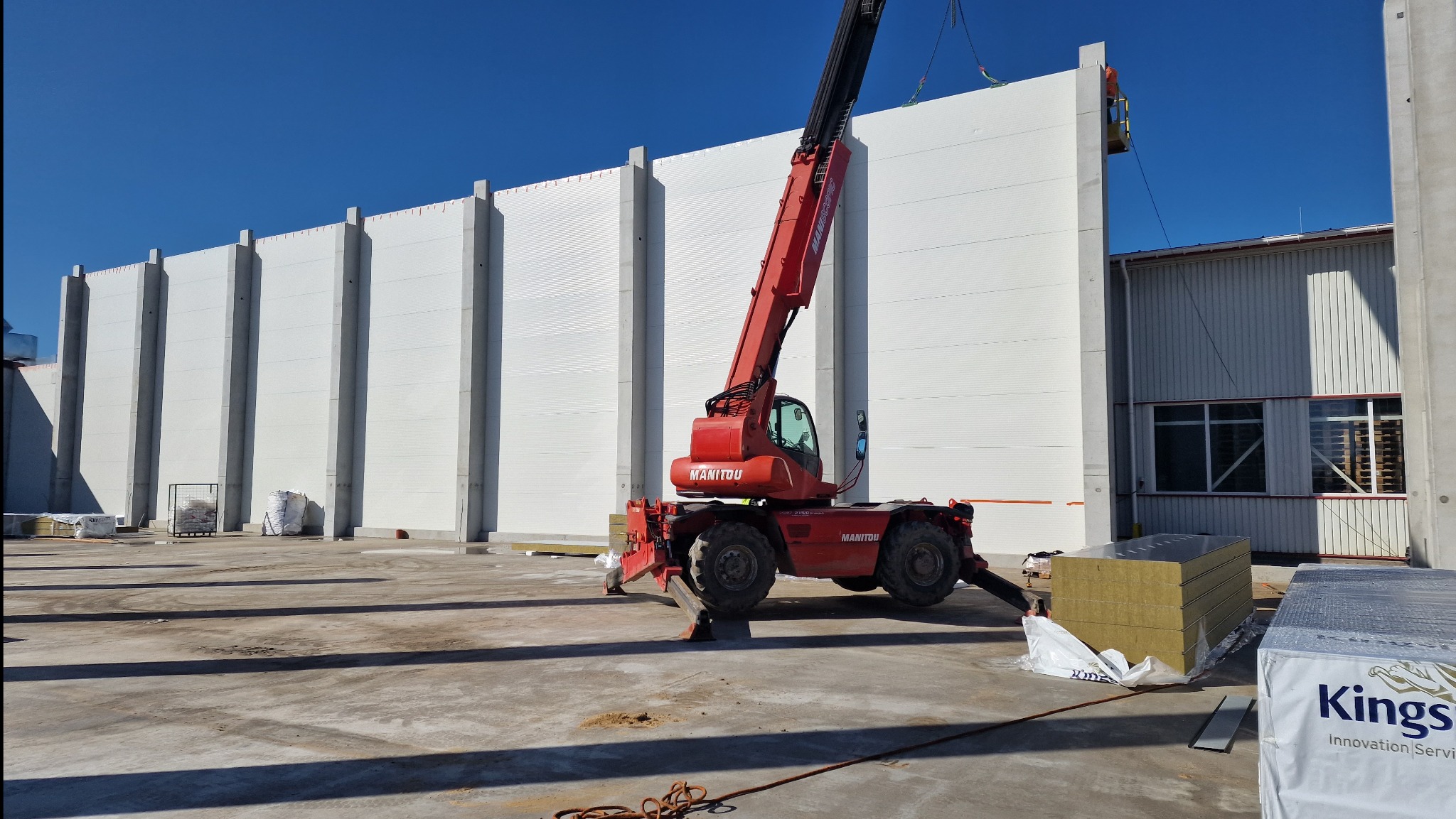Montaż białych płyt warstwowych na hali przemysłowej za pomocą czerwonego podnośnika Manitou. Widoczne materiały izolacyjne Kingspan i pracownik na podnośniku koszowym.
