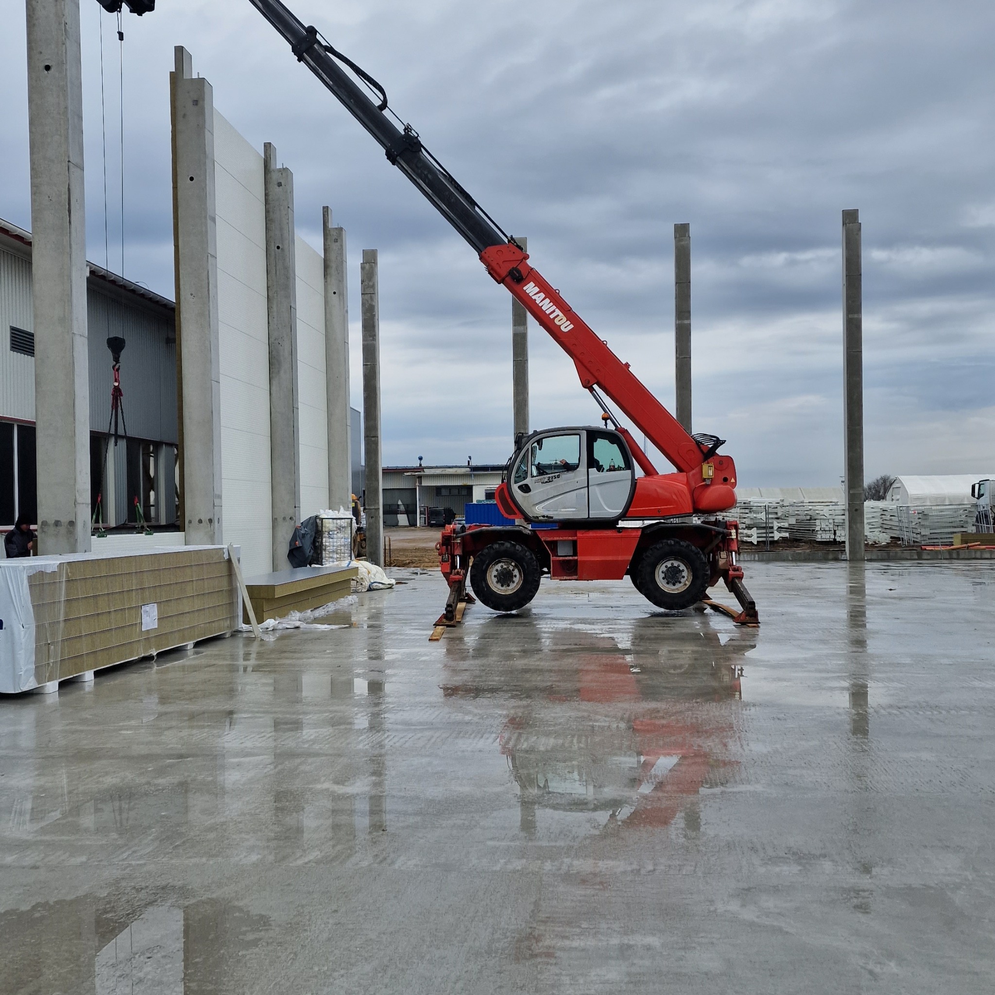 Montaż płyt warstwowych na konstrukcji hali z użyciem czerwonego podnośnika Manitou. Widoczne kolumny i przygotowane płyty. Mokra posadzka odbija elementy konstrukcji.