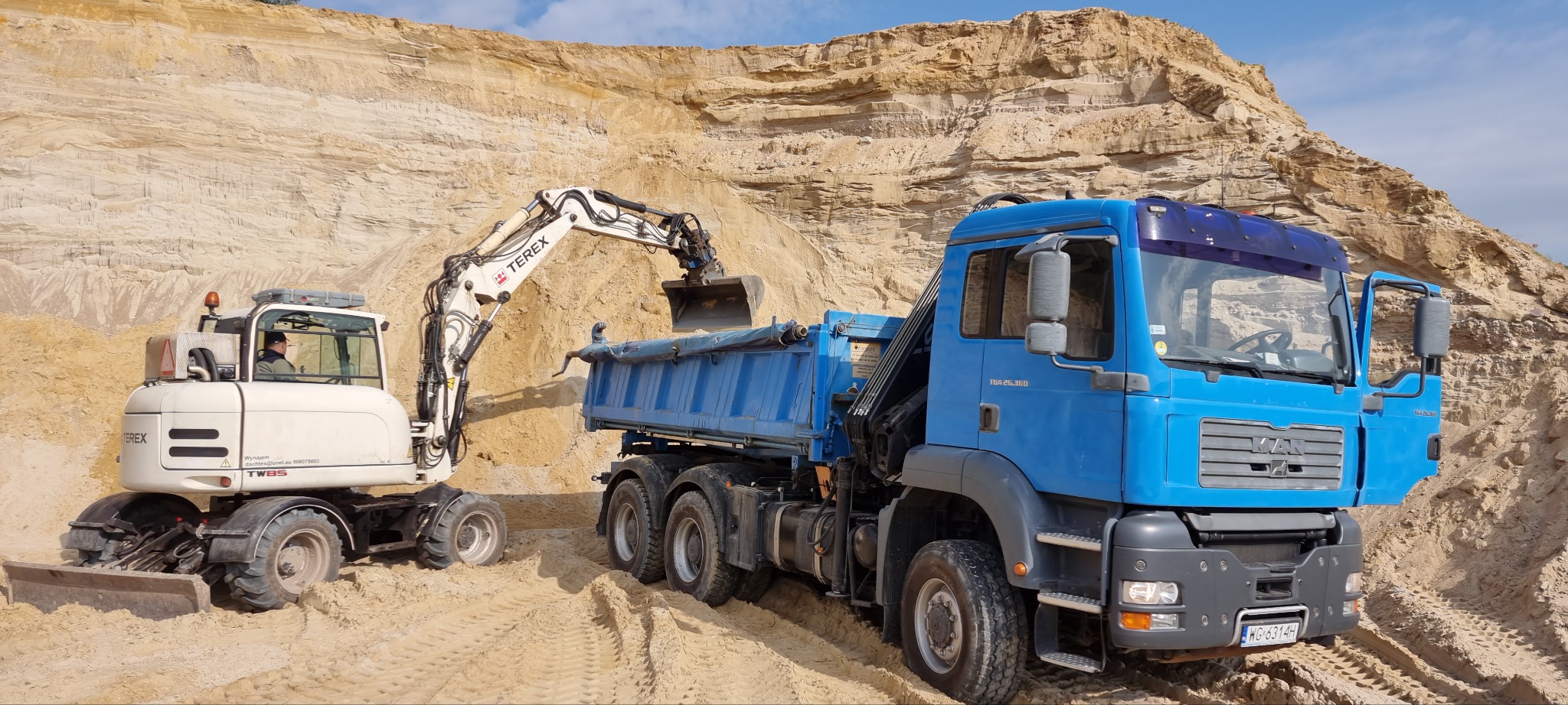 Błękitna ciężarówka MAN i biała koparka Terex ładują piasek na tle piaszczystego wzgórza. Widoczny operator koparki i tablica rejestracyjna ciężarówki.