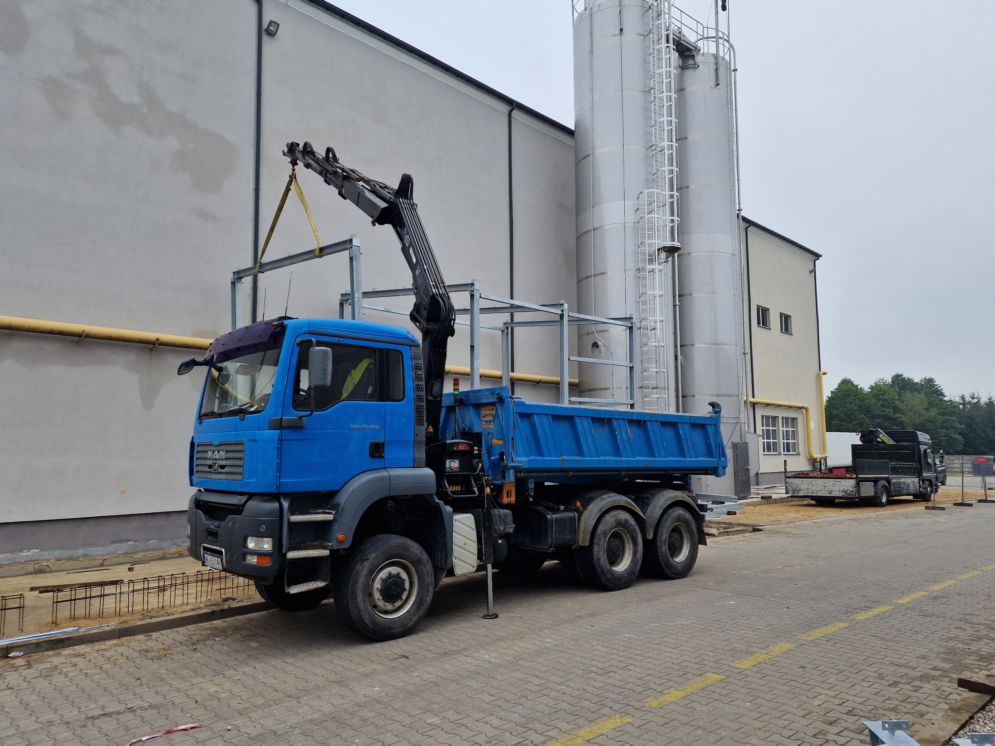 Niebieska ciężarówka z dźwigiem podnosi element konstrukcji stalowej na tle budynku przemysłowego z silosami. Widoczne zbrojenia i kostka brukowa.
