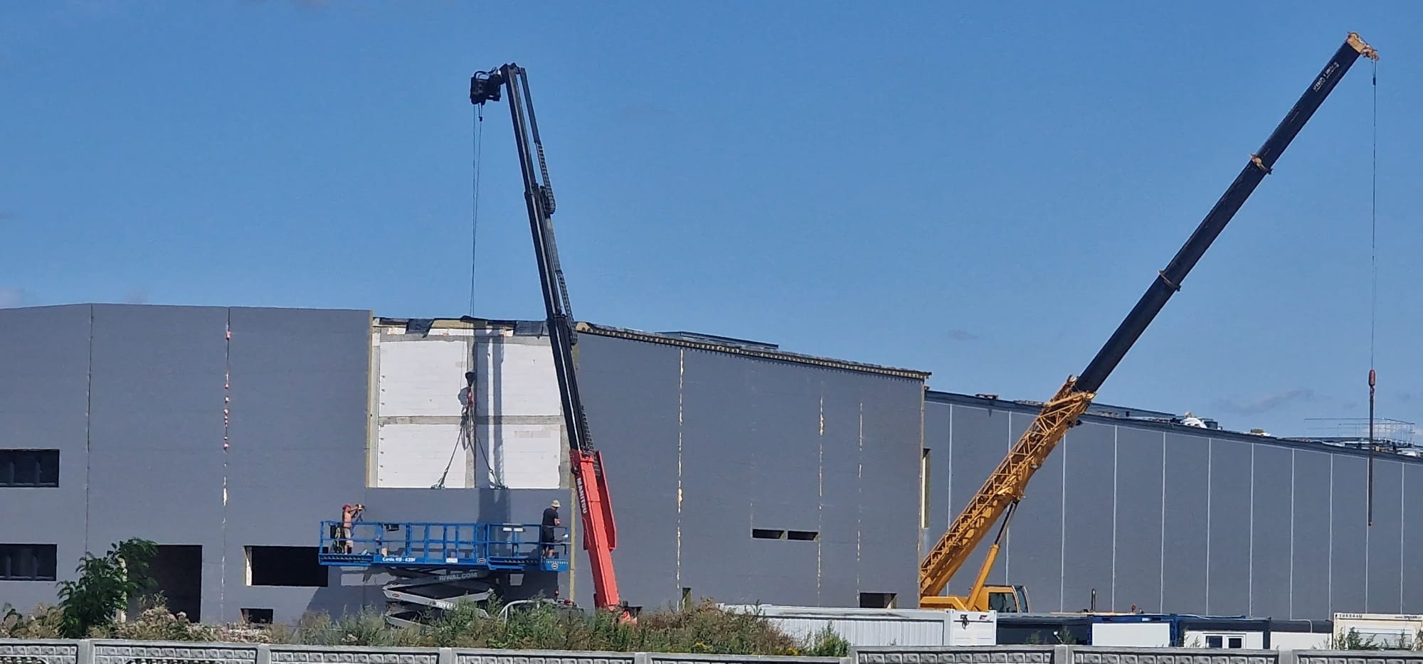 Montaż płyty warstwowej na hali przemysłowej z użyciem dźwigu i podnośnika koszowego. Pracownicy w kaskach. Widok na budynek w trakcie prac.