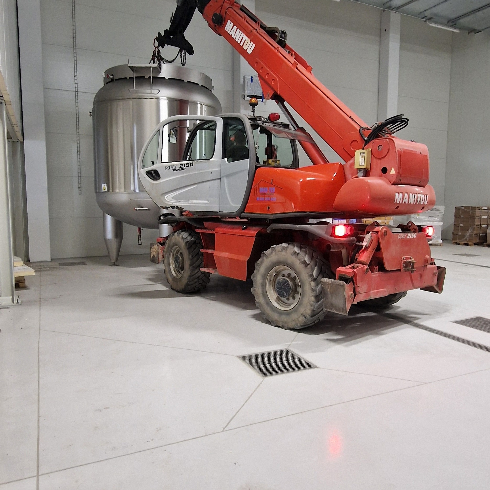 Czerwony podnośnik Manitou transportuje duży, metalowy zbiornik wewnątrz hali w Sobolewie. Maszyna stoi na betonowej posadzce z kratkami odpływowymi.