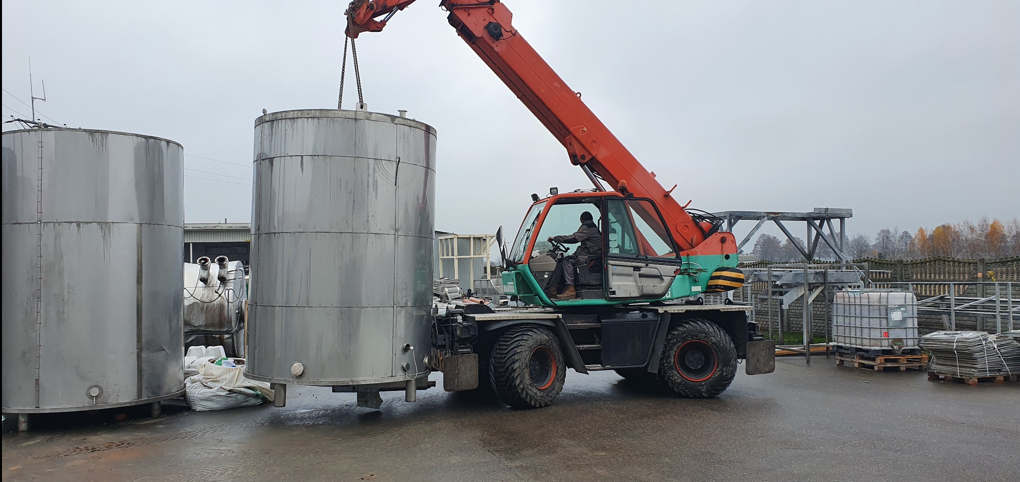 Pomarańczowo-zielona ładowarka teleskopowa podnosi srebrny zbiornik na zewnątrz w pochmurny dzień. Widoczne inne zbiorniki i elementy konstrukcyjne.