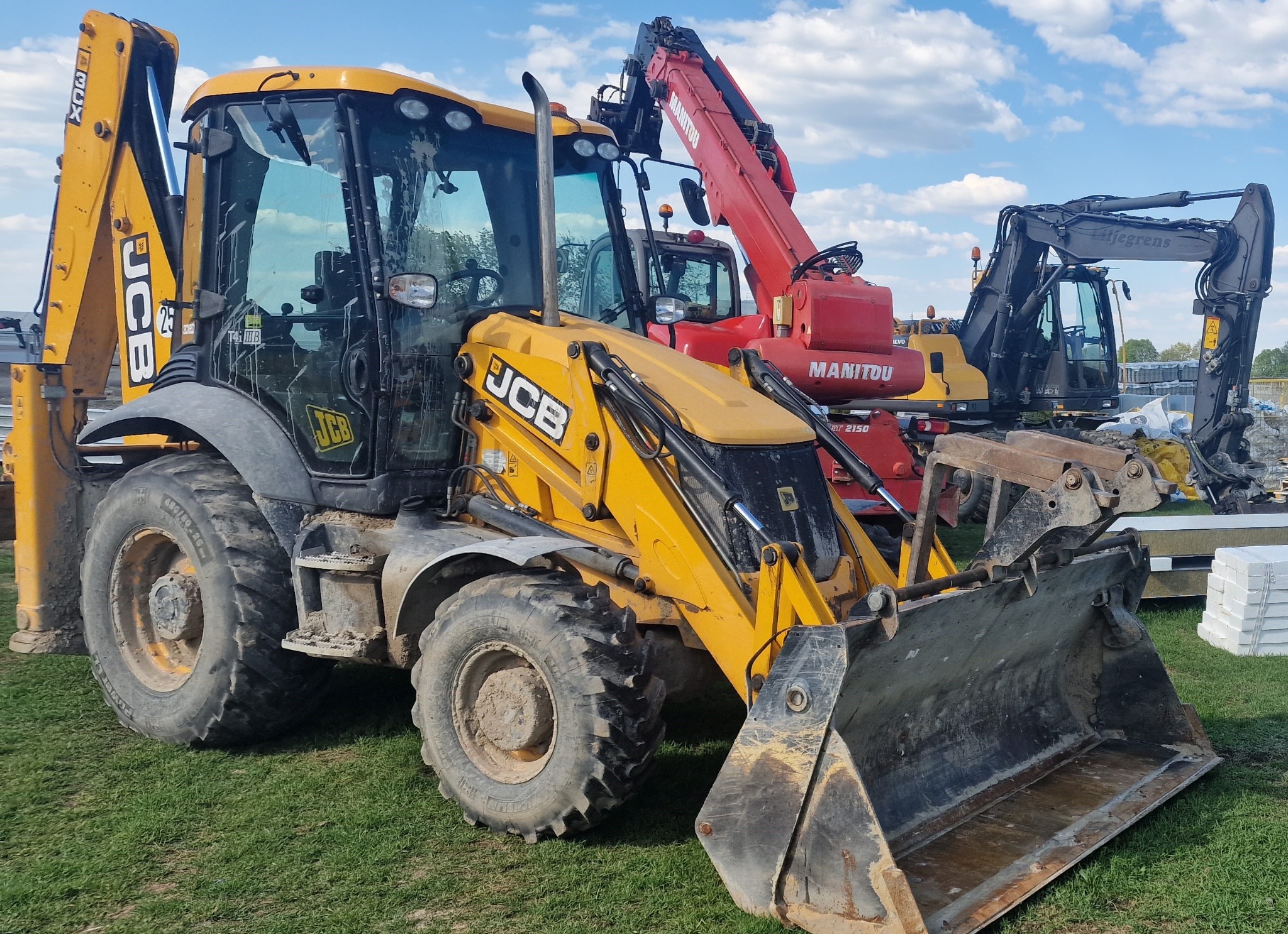 Żółta koparko-ładowarka JCB na trawiastym terenie, z widocznymi śladami użytkowania, na tle innych maszyn budowlanych i błękitnego nieba z chmurami.
