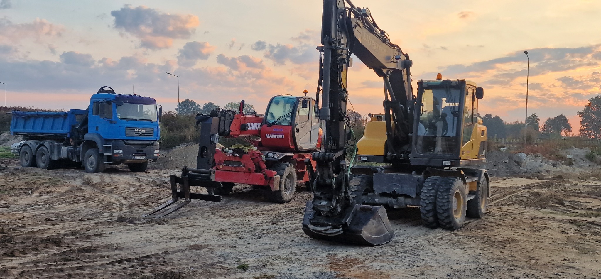 Żółta koparka kołowa, czerwony podnośnik teleskopowy Manitou i niebieska wywrotka MAN na placu budowy o zmierzchu. Widoczne ślady opon na piasku.