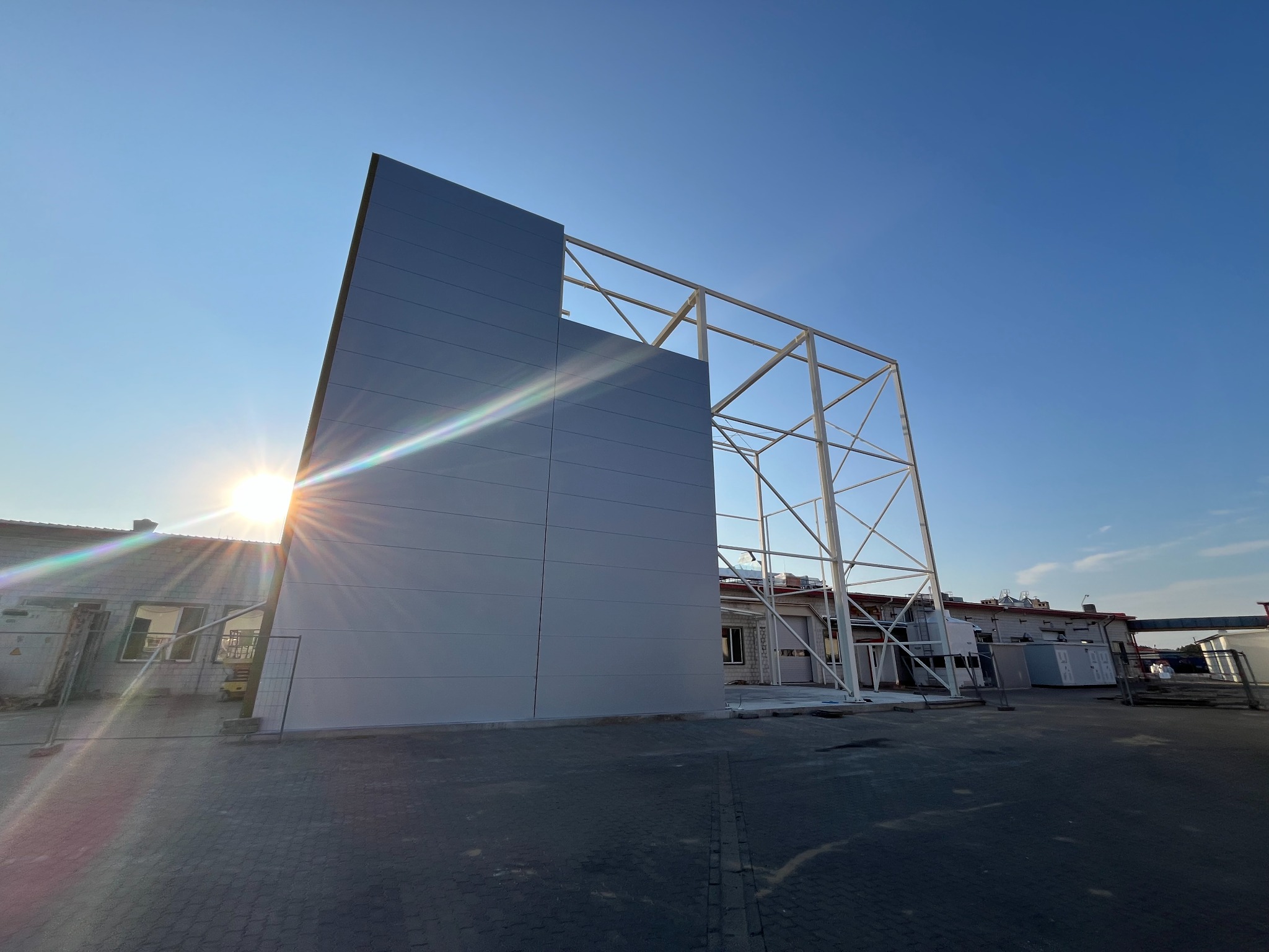 Wznoszona hala z płyt warstwowych w blasku słońca. Widoczna stalowa konstrukcja i fragmenty zamontowanych paneli. Częściowo ukończony budynek w tle.
