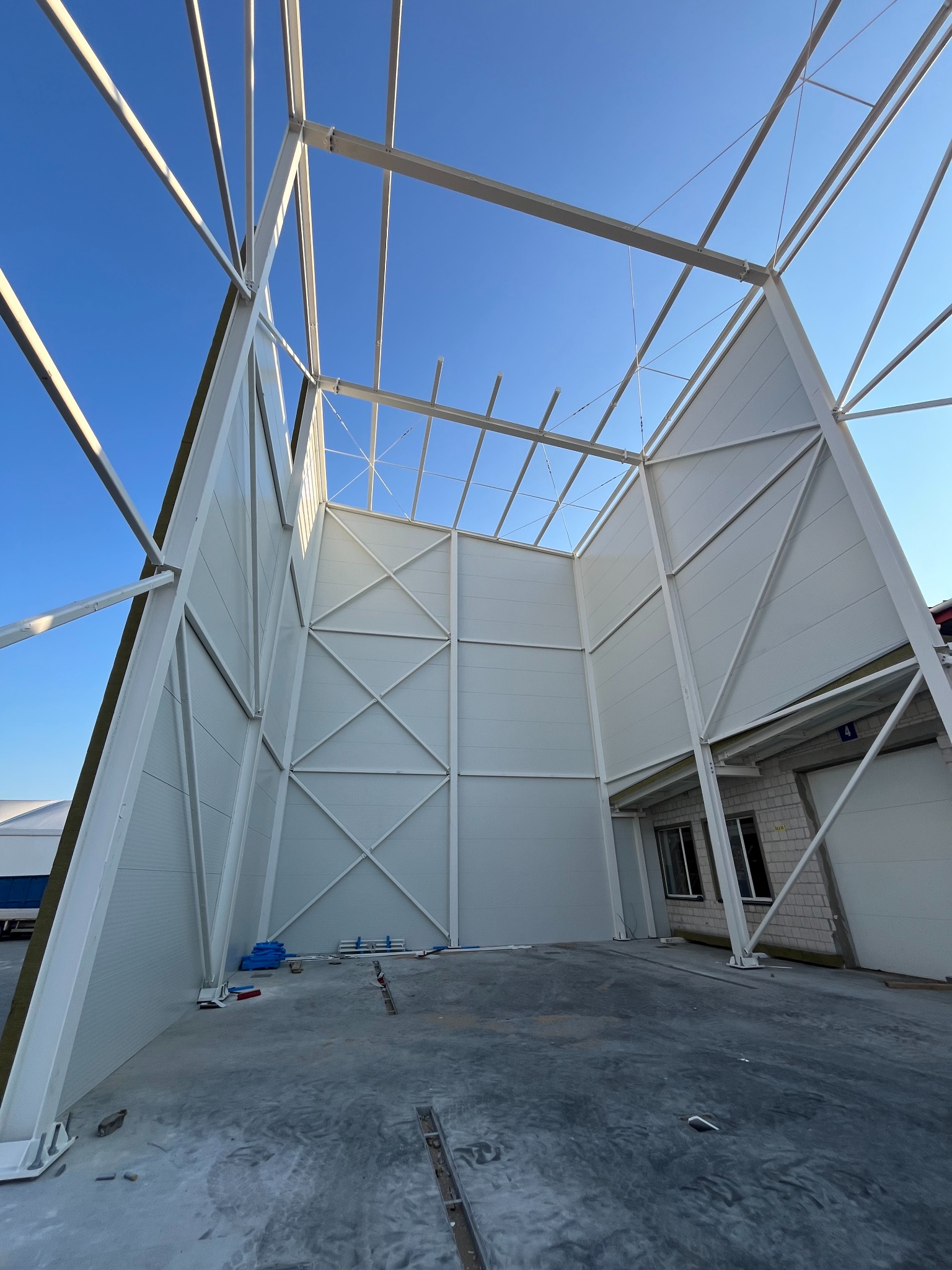 Wnętrze hali w trakcie budowy: białe ściany z płyt warstwowych, stalowa konstrukcja nośna, widok na niebo, betonowa posadzka. Ujęcie z dołu, szeroki kadr.