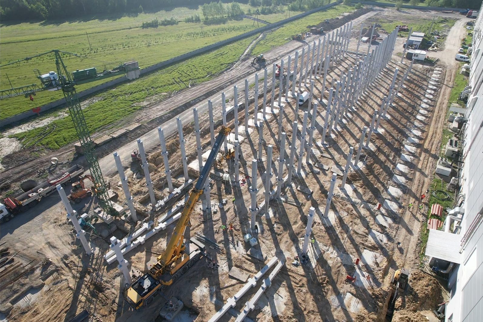 Budowa hali: widok z lotu ptaka na konstrukcję z betonowych słupów, żuraw i sprzęt budowlany na placu. W tle zielone pola i las.