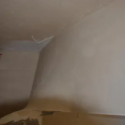 Świeżo otynkowana ściana wewnętrzna z widocznymi instalacjami hydraulicznymi, sufit z wystającym kablem elektrycznym, widoczne niedoskonałości wykończenia.