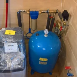 Zestaw urządzeń do uzdatniania wody i hydrofor w pomieszczeniu wyłożonym beżowymi płytkami, widoczne rury i zawory.