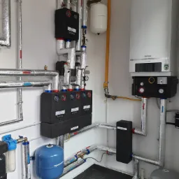 Nowoczesna kotłownia z piecem gazowym Buderus, rozdzielaczami, grupami pompowymi, rurami w izolacji termicznej oraz zbiornikami wyrównawczymi, zainstalowana w pomieszczeniu z białymi ścianami...