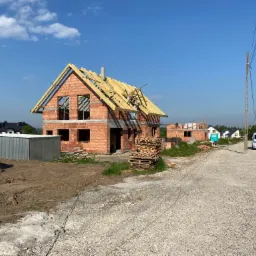 Niedokończony dom jednorodzinny z czerwonej cegły z drewnianą konstrukcją dachu, widoczny stos palet z cegłami oraz droga dojazdowa z białym samochodem dostawczym w tle.