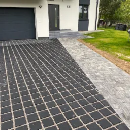 Nowoczesny dom jednorodzinny z antracytową bramą garażową i drzwiami wejściowymi, z dwoma rodzajami nawierzchni z kostki brukowej: czarnej ażurowej i szarej płaskiej, prowadzącymi do wejścia.