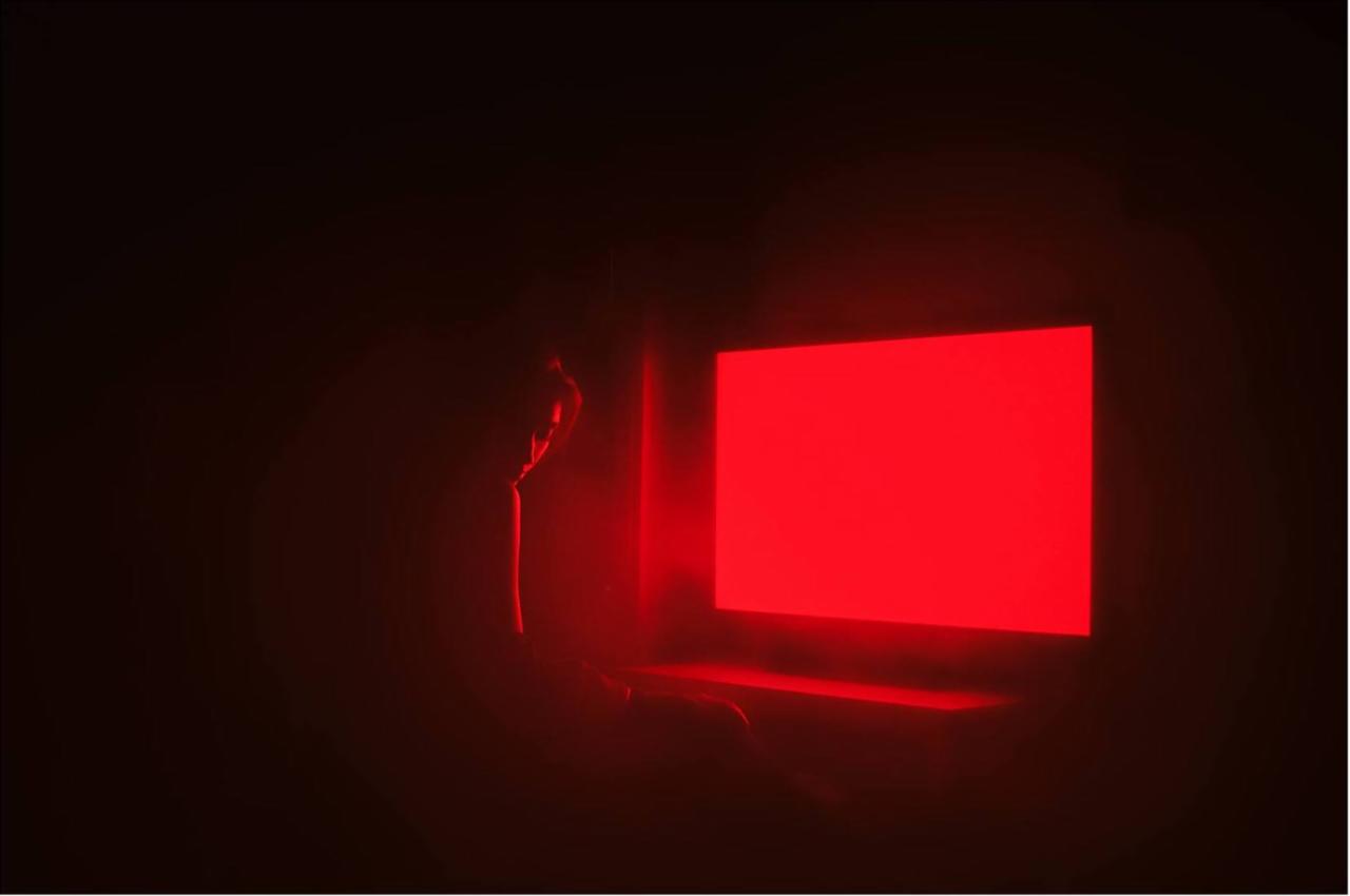Sylwetka kobiety w czerwonym świetle, siedzącej przed jasnym, prostokątnym ekranem, tworząca minimalistyczną kompozycję.