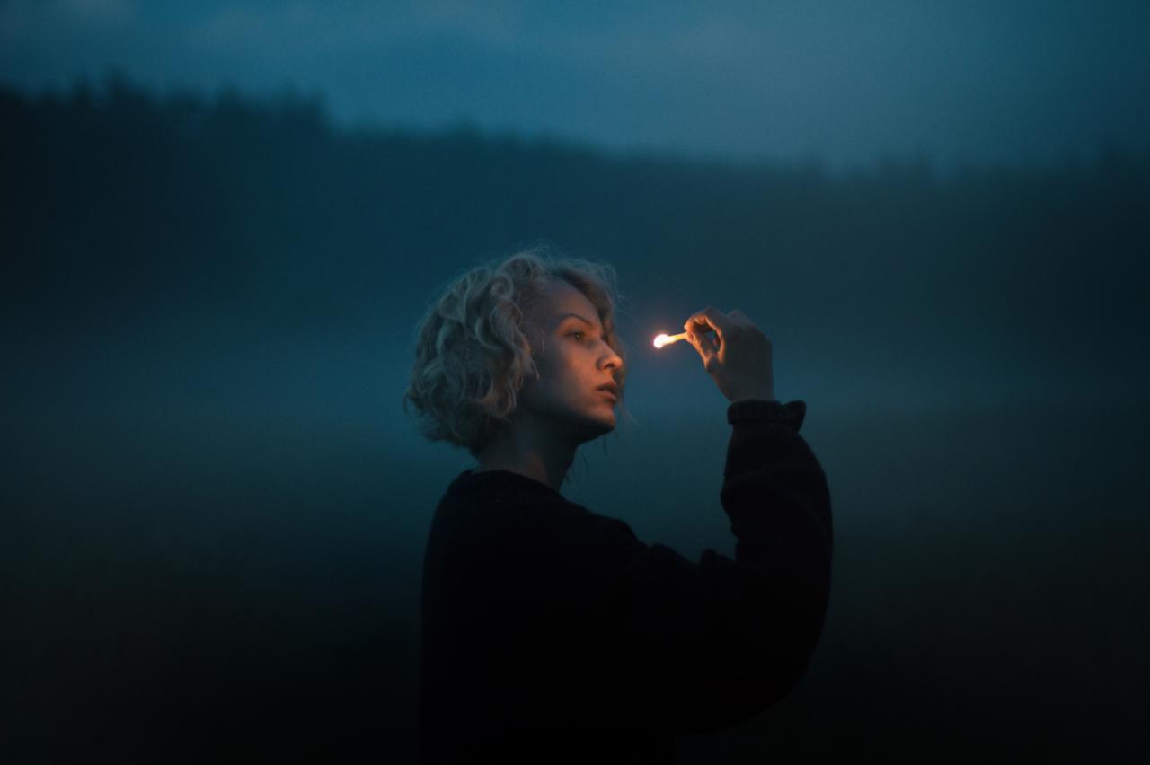 Kobieta z kręconymi blond włosami trzyma zapaloną zapałkę na ciemnym tle, oświetlona delikatnym blaskiem płomienia.