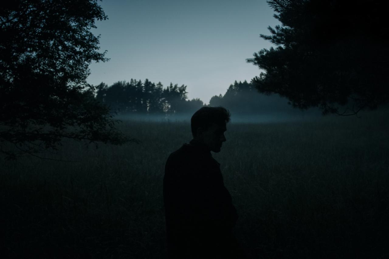 Sylwetka mężczyzny na tle zalesionego krajobrazu spowitego mgłą, w chłodnej tonacji kolorystycznej.