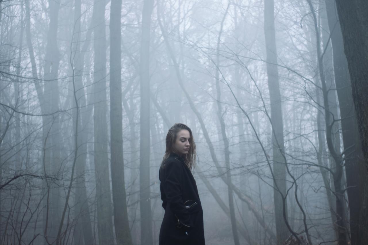 Kobieta w czarnym płaszczu w mglistym lesie, pochylona głowa, melancholijny nastrój, drzewa bez liści w tle.