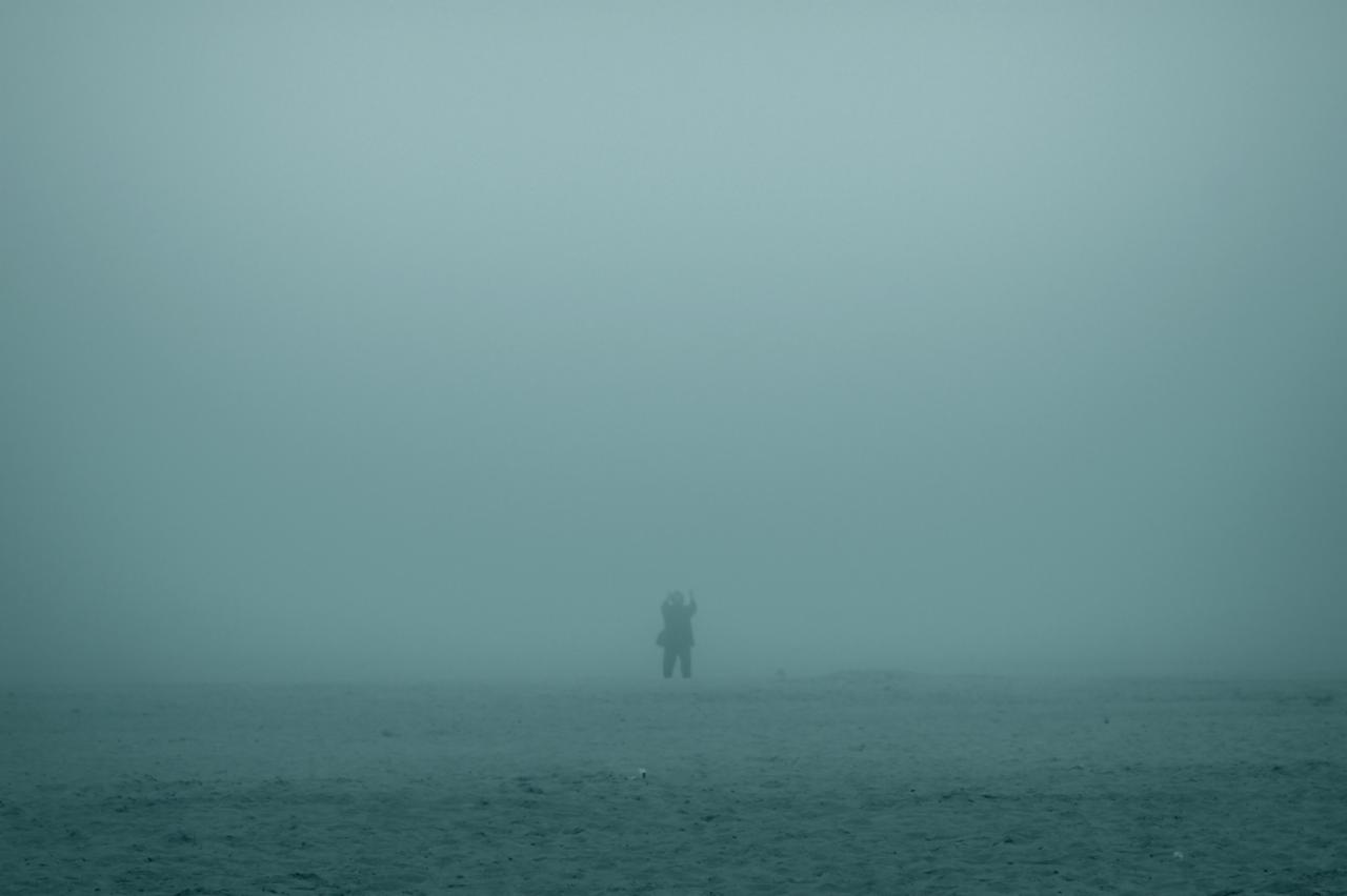 Sylwetka osoby w oddali na tle gęstej, turkusowej mgły nad ciemnym, nierównym terenem.