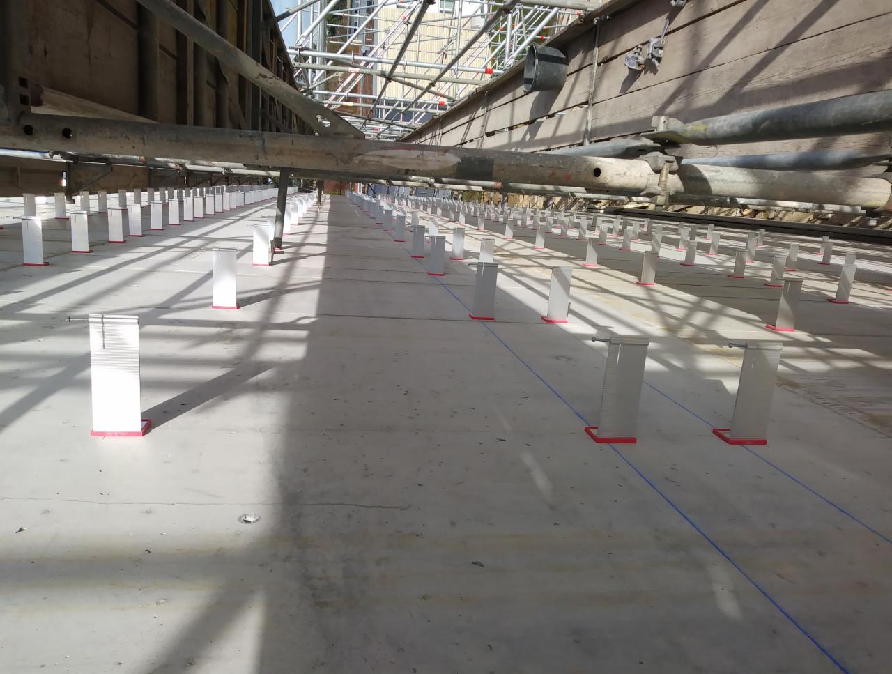 Przygotowanie podłoża: rzędy białych podpór z czerwonymi podstawami, ustawione równo na betonowej powierzchni, wyznaczające linie pod przyszłą konstrukcję, widok spod rusztowania.