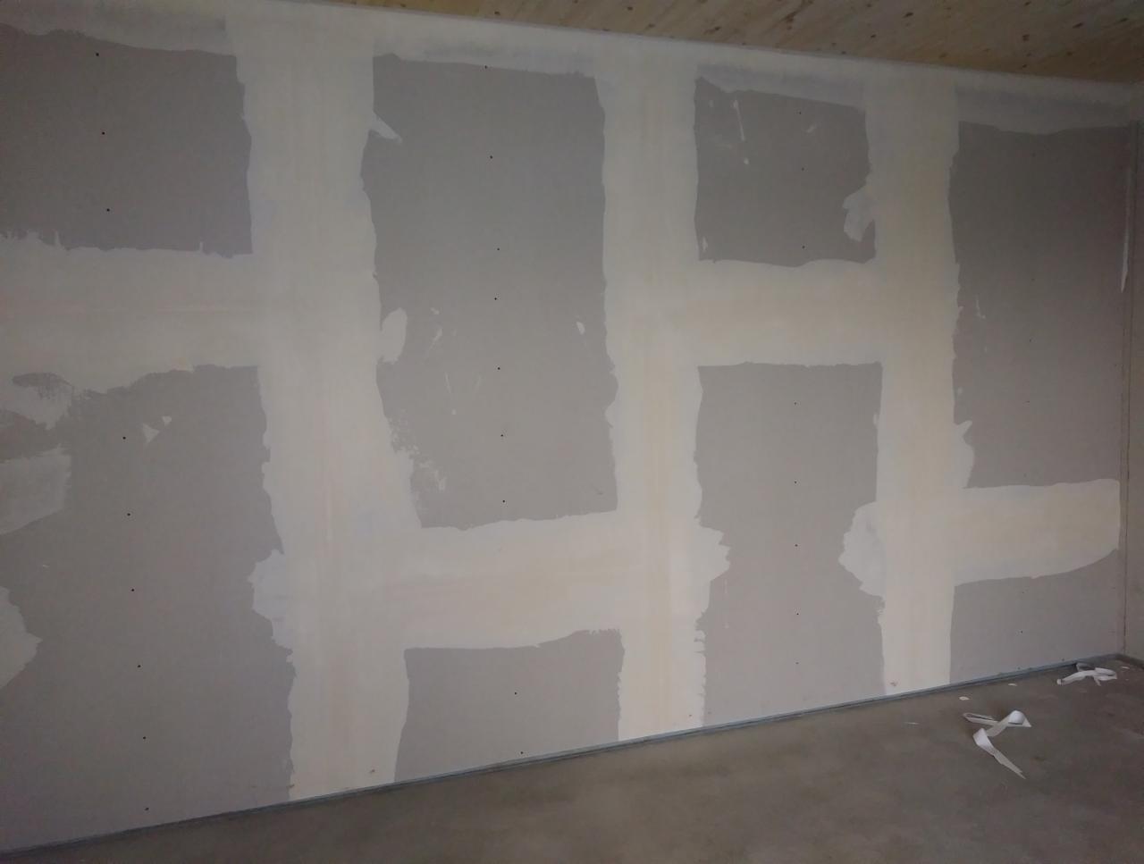 Ściana z płyt gipsowo-kartonowych w trakcie łączenia, widoczne paski wzmacniające na łączeniach, surowy beton na podłodze, drewniany sufit.