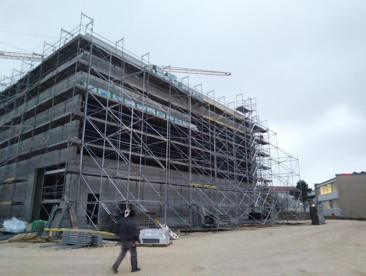 Budynek w trakcie budowy, pokryty rusztowaniem, widoczny żuraw w tle, pochmurne niebo.
