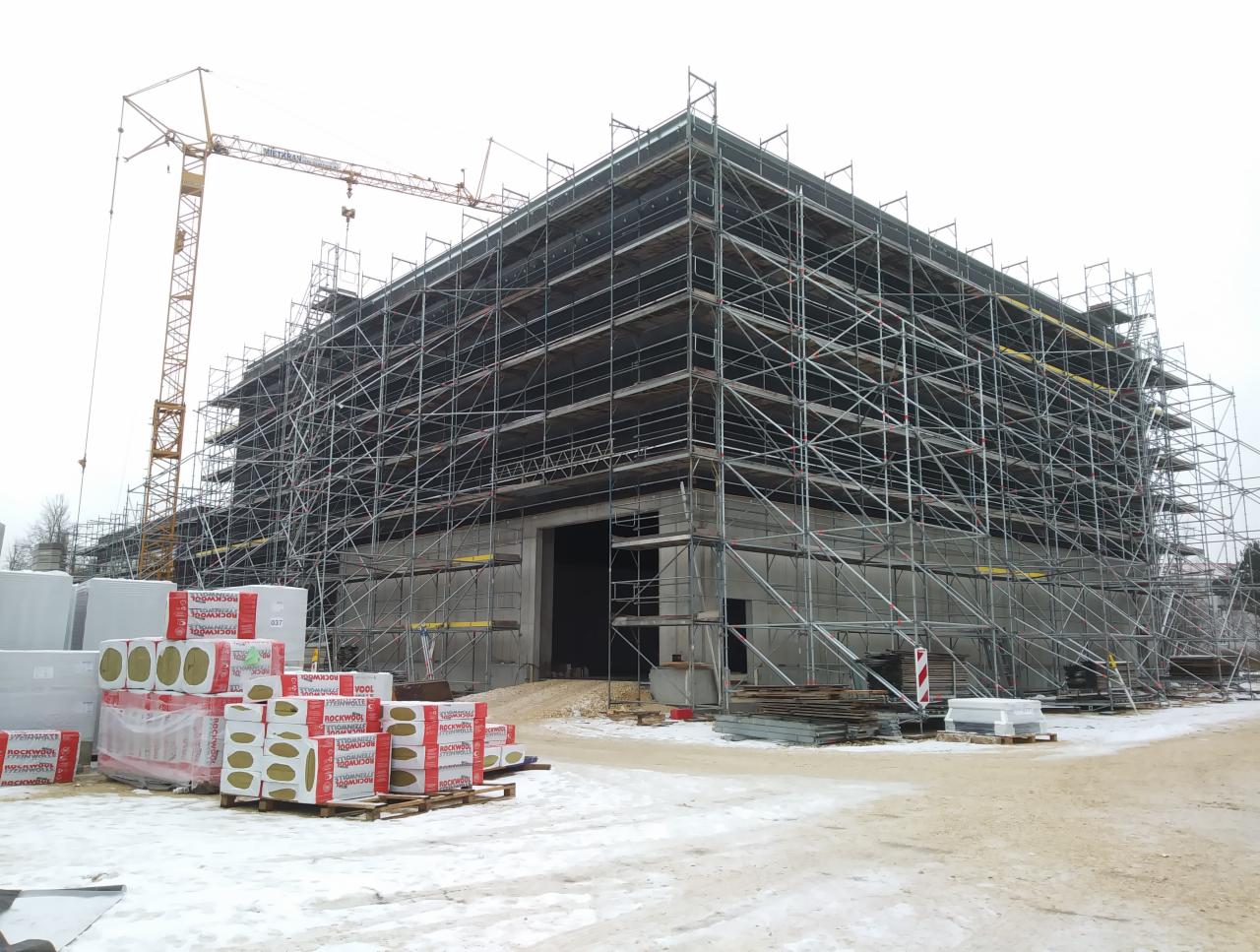 Budynek w trakcie ocieplania, otoczony rusztowaniami, z widocznym dźwigiem i materiałami izolacyjnymi Rockwool na paletach.