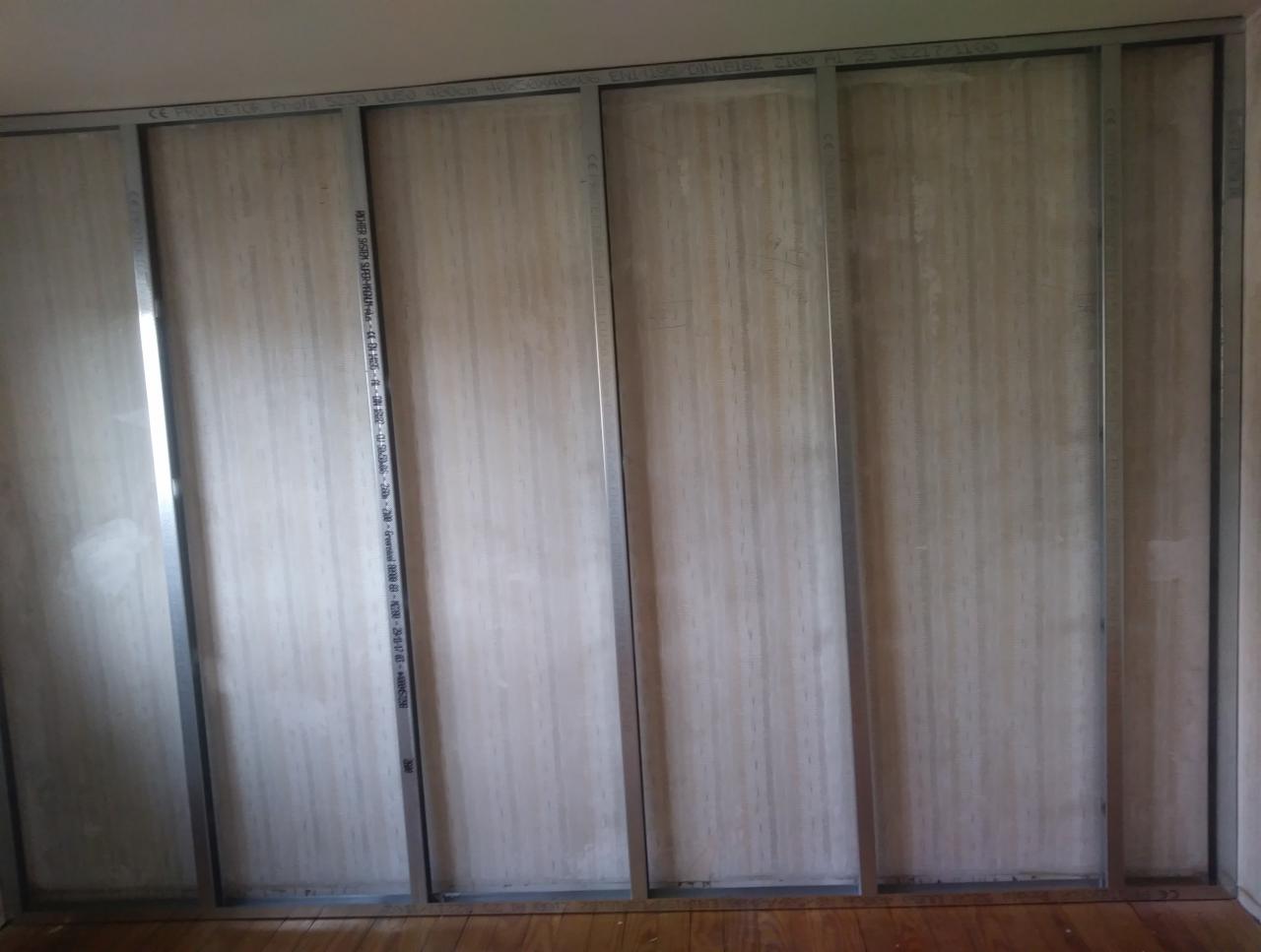 Konstrukcja ściany działowej z płyt kartonowo-gipsowych na metalowym stelażu, widoczne płyty częściowo zamontowane, drewniana podłoga.