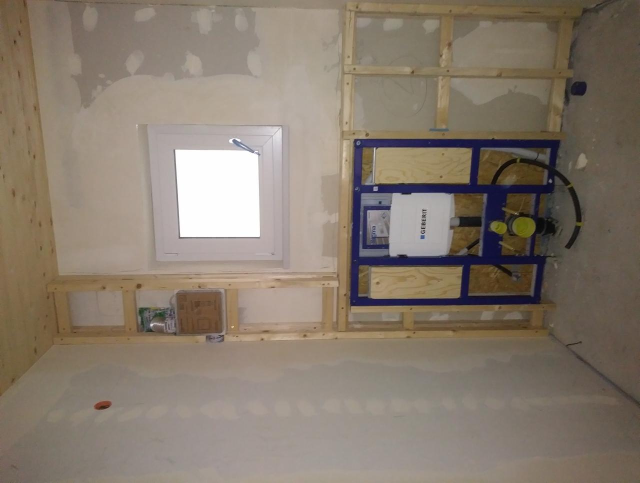 Ściana w trakcie remontu, z zamontowanym stelażem podtynkowym WC marki Geberit, obudowana drewnianą konstrukcją, częściowo oszpachlowana, z widocznym oknem i materiałami budowlanymi.