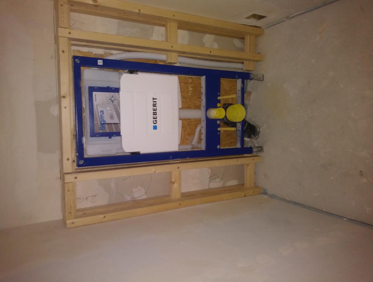 Stelaż podtynkowy Geberit Sigma zamontowany w drewnianej konstrukcji, widoczne rury i elementy instalacji w surowym stanie, przygotowanie do obudowy płyty gipsowo-kartonowej.