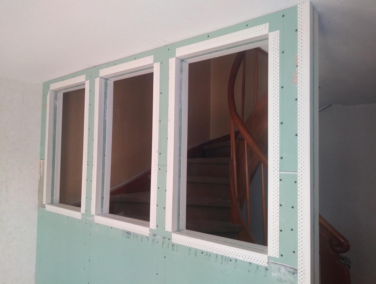 Montaż płyty gipsowo-kartonowej z metalowymi narożnikami wokół trzech otworów okiennych, widoczne schody w tle.