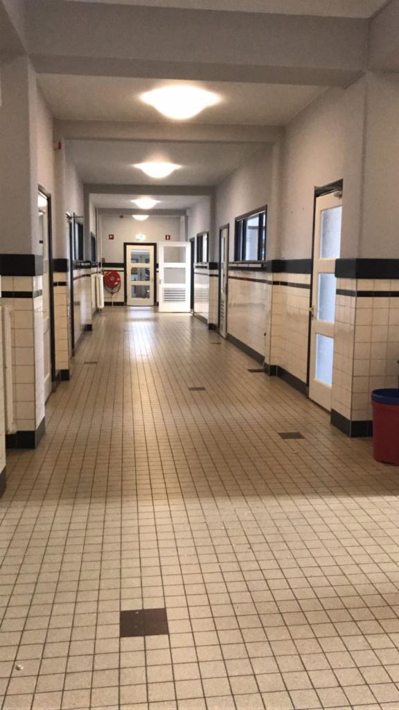 Długi, korytarz w budynku z białymi, kwadratowymi płytkami na ścianach do połowy wysokości i na podłodze, z czarnymi akcentami na krawędziach ścian i podłogi, białe drzwi po obu stronach korytarza,...