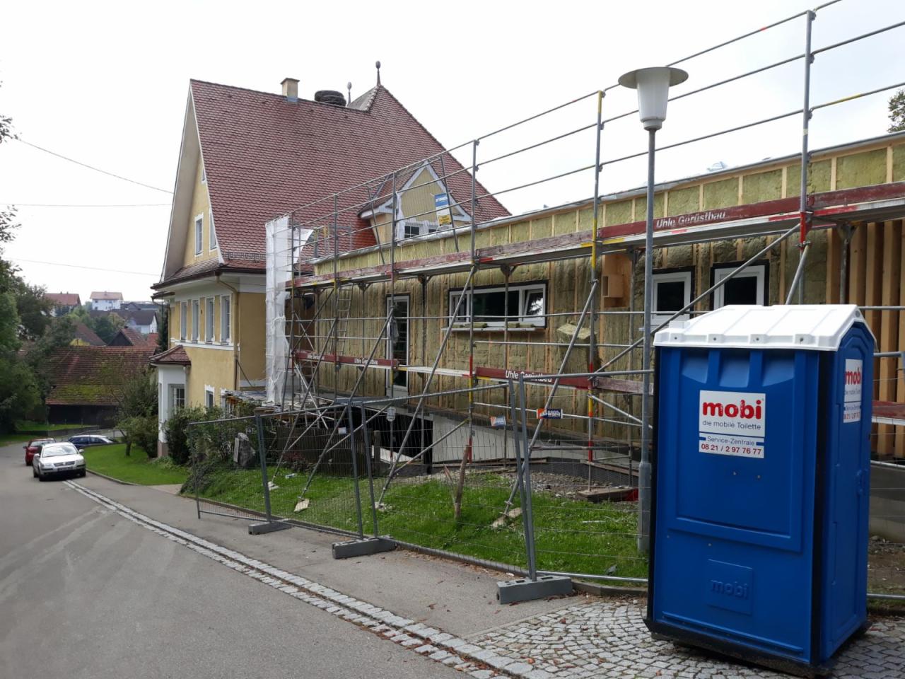 Dom w trakcie docieplania, widoczne rusztowania, warstwa izolacji na ścianach, okna, oraz przenośna toaleta na pierwszym planie.