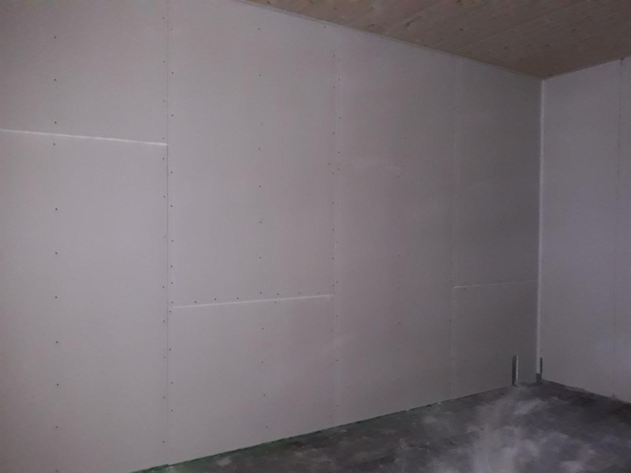 Wnętrze pokoju w trakcie remontu, ściany wyłożone surowymi płytami gipsowo-kartonowymi, widoczne wkręty mocujące, sufit drewniany, podłoga betonowa.