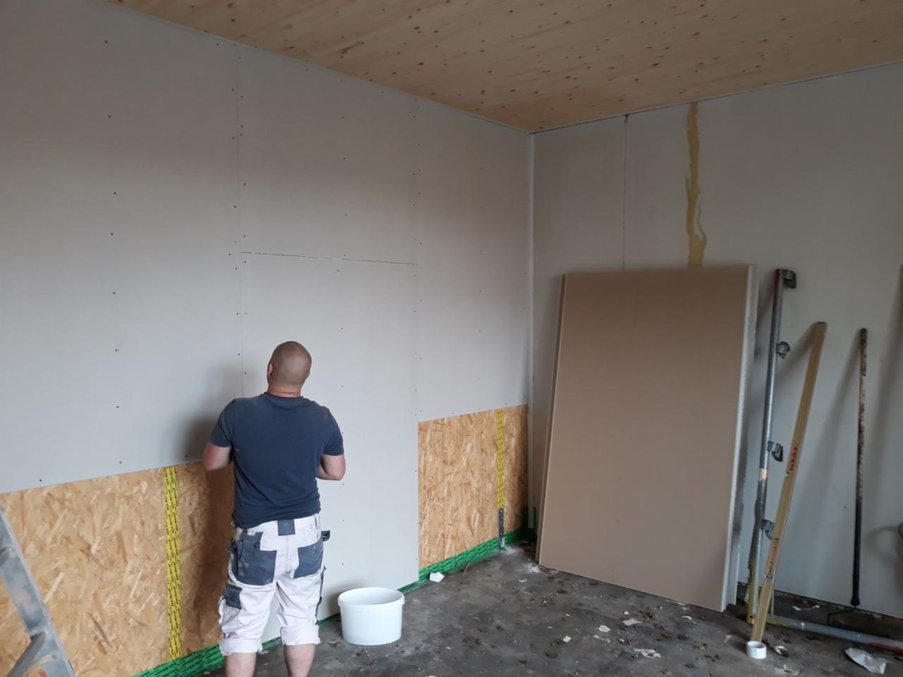 Instalacja płyty gipsowo-kartonowej na ścianie, widoczny pracownik w roboczych spodniach, fragment drewnianego sufitu i podkład z płyty OSB zabezpieczony folią.