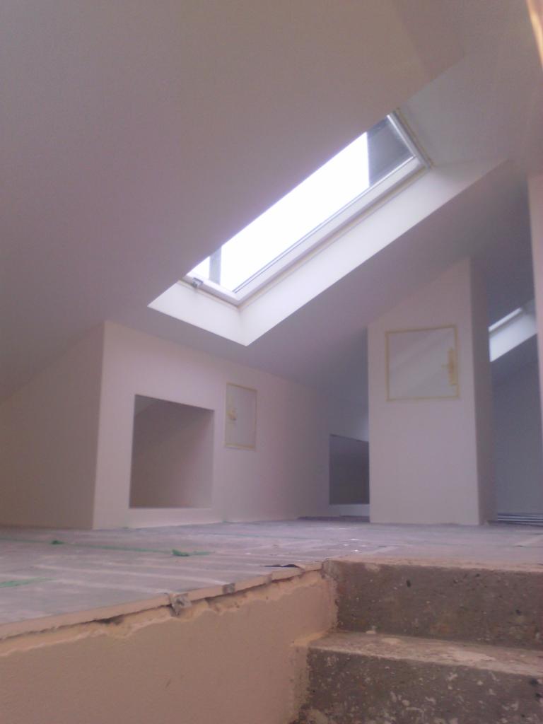 Wnętrze poddasza w trakcie wykańczania, widoczne okno dachowe, ściany po wstępnym wyrównaniu, przygotowane do dalszych prac, zarys schodów w dolnym rogu kadru.