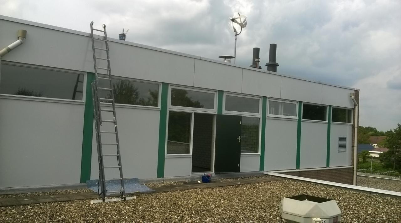 Budynek przemysłowy z drabina opartą o ścianę, widoczne ślady po malowaniu, otwarte drzwi i wiadro z farbą na dachu pokrytym żwirem.