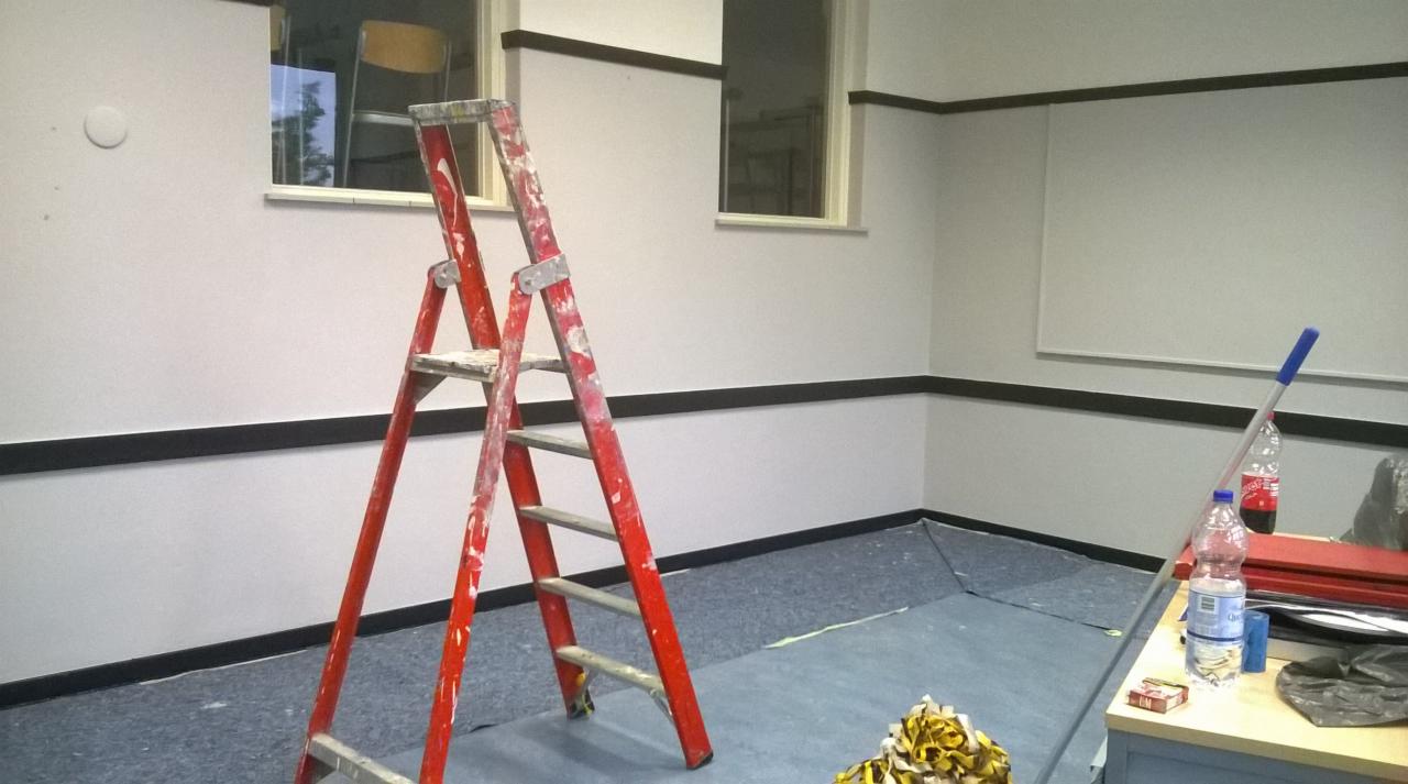 Wnętrze pokoju po odświeżeniu, z pomalowanymi ścianami na biało i ciemnym pasem w dolnej części, widoczna czerwona drabina malarska, butelki z napojami i inne akcesoria malarskie.