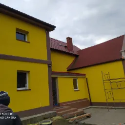 Żółta elewacja budynku z czerwoną dachówką, widoczne rusztowanie i osoba w czapce, sugerujące prace wykończeniowe.