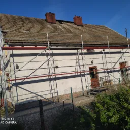 Dom jednorodzinny z brązowym dachem i białymi ścianami, otoczony rusztowaniem; widoczne dwa kominy, antena i zarośla w ogrodzie.