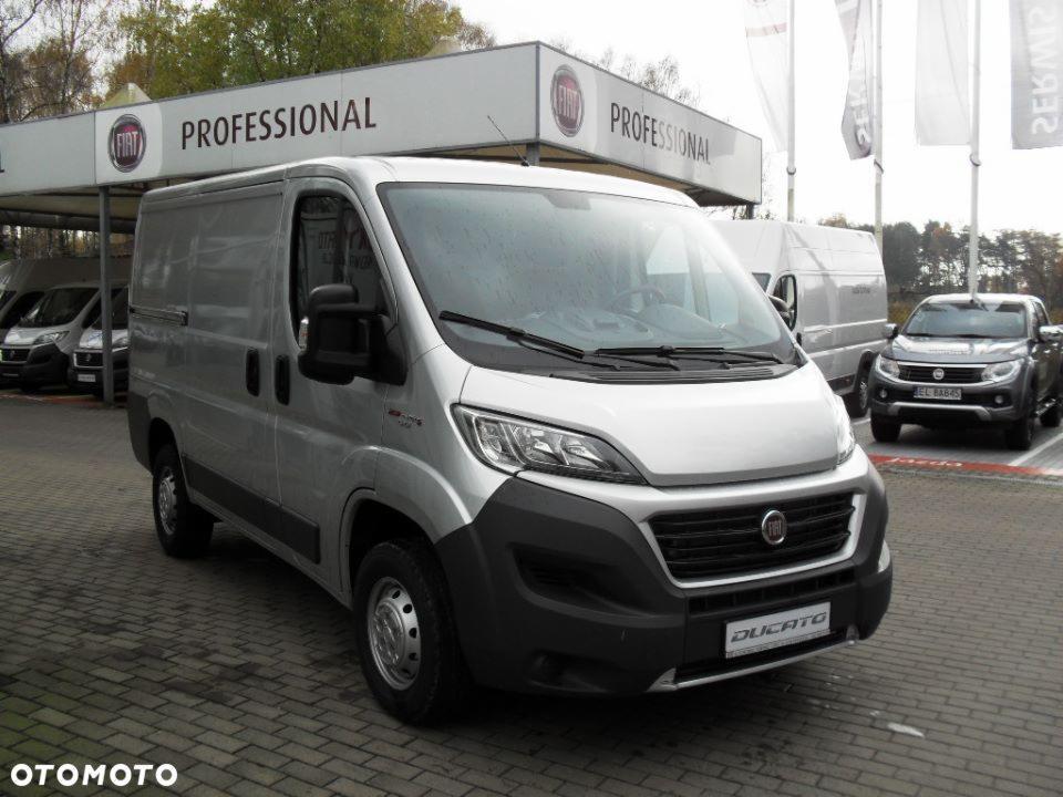 Srebrny samochód dostawczy Fiat Ducato Professional na placu przed salonem, widoczne inne pojazdy w tle.