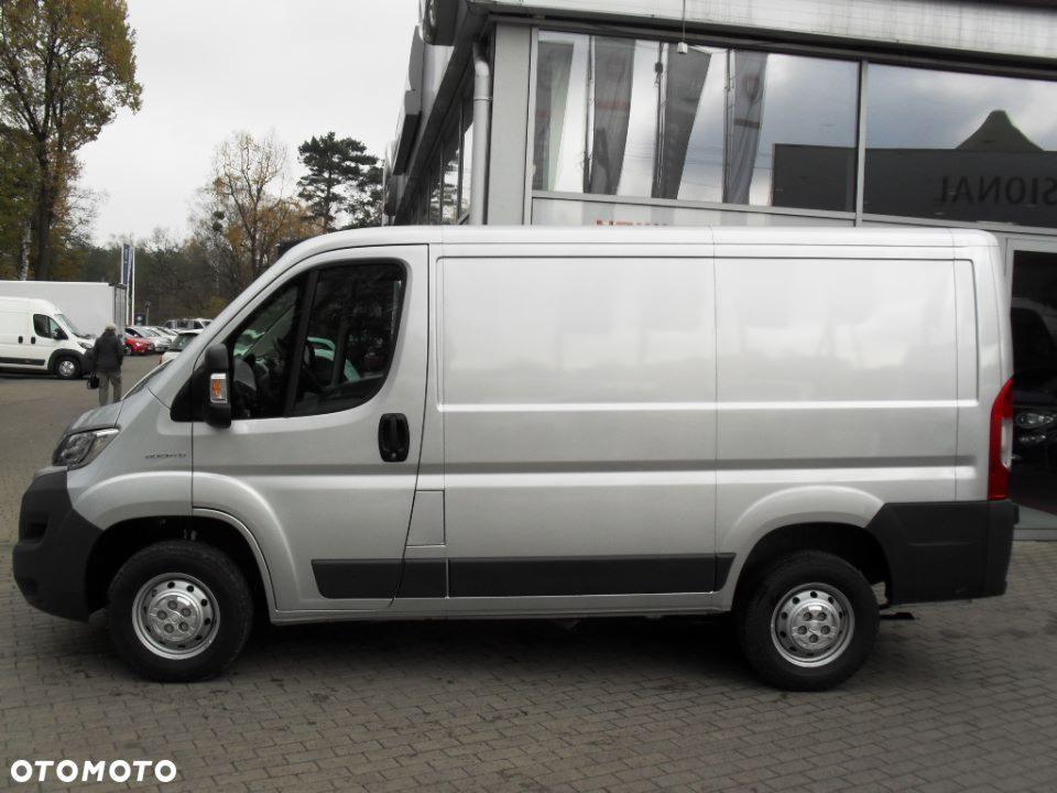 Srebrny, dostawczy bus marki Fiat Ducato, zaparkowany na brukowanej nawierzchni przed przeszklonym budynkiem salonu samochodowego, widok z boku.