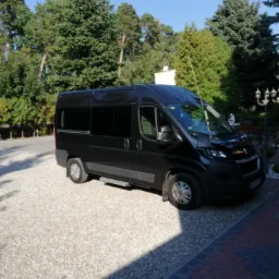 Czarny bus z przyciemnianymi szybami, zaparkowany na żwirowym podjeździe, otoczony zielenią drzew.