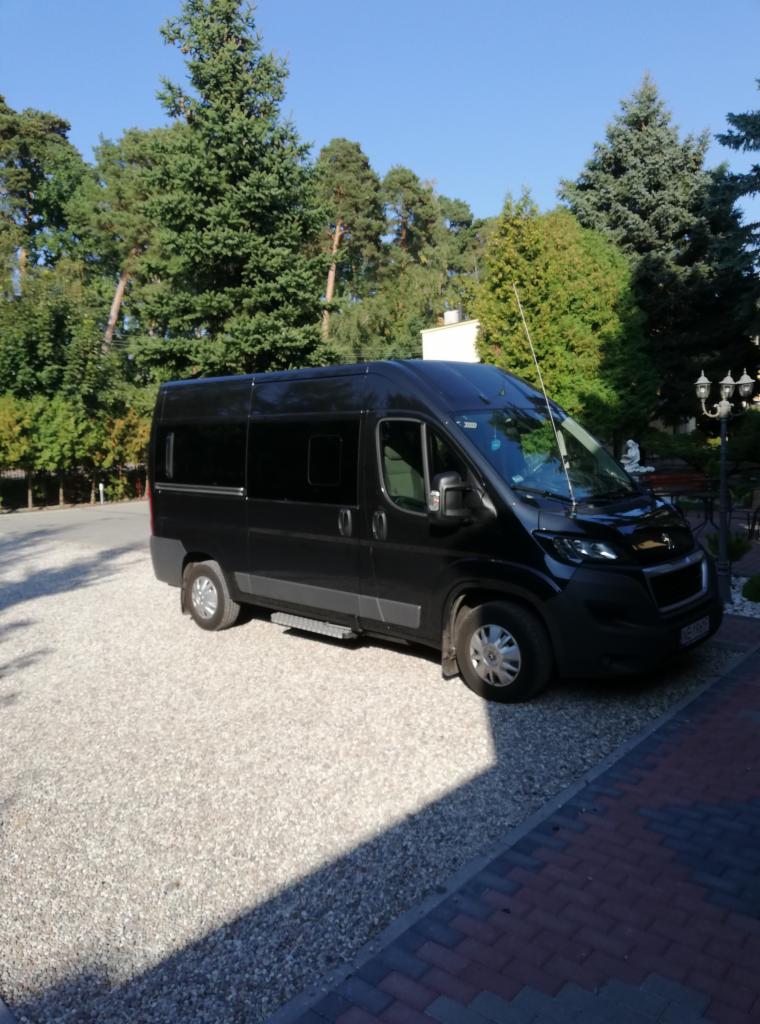 Czarny bus z przyciemnianymi szybami, zaparkowany na żwirowym podjeździe, otoczony zielenią drzew.