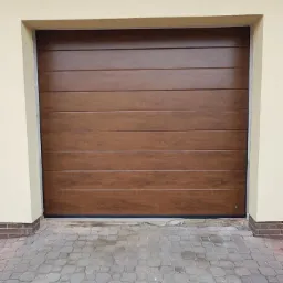 Zamknięta, segmentowa brama garażowa w kolorze drewna, z poziomymi panelami, zamontowana w beżowej elewacji budynku i wyłożona kostką brukową.