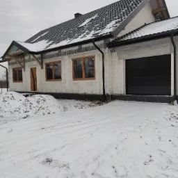 Surowy stan domu jednorodzinnego w zimowej scenerii; widoczne okna z drewnianymi ramami i czarna brama garażowa, ściany z bloków, część dachu pokryta dachówką, śnieg na ziemi.