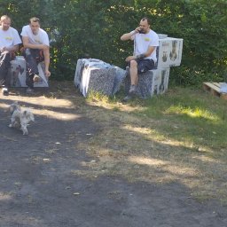 Jankes-Bud Janusz Smolen - Trzech mężczyzn w roboczych ubraniach odpoczywa na kostkach izolacyjnych z polistyrenu ekstrudowanego, mały pies biegnie po ziemi, w tle biały samochód i zielona roślinność.