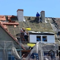 Jankes-Bud Janusz Smolen - Ekipa dekarzy na dachu podczas wymiany pokrycia, widoczne łatkowanie, kominy i częściowo ułożona dachówka ceramiczna, zabezpieczenia rusztowań w dolnej części kadru.