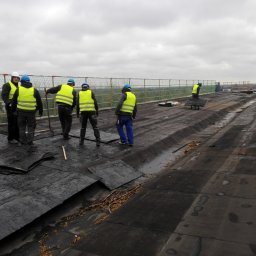Jankes-Bud Janusz Smolen - Ekipa dekarzy w kamizelkach odblaskowych usuwa starą papę z płaskiego dachu budynku, zabezpieczonego siatką ochronną, widok na miasto w tle.